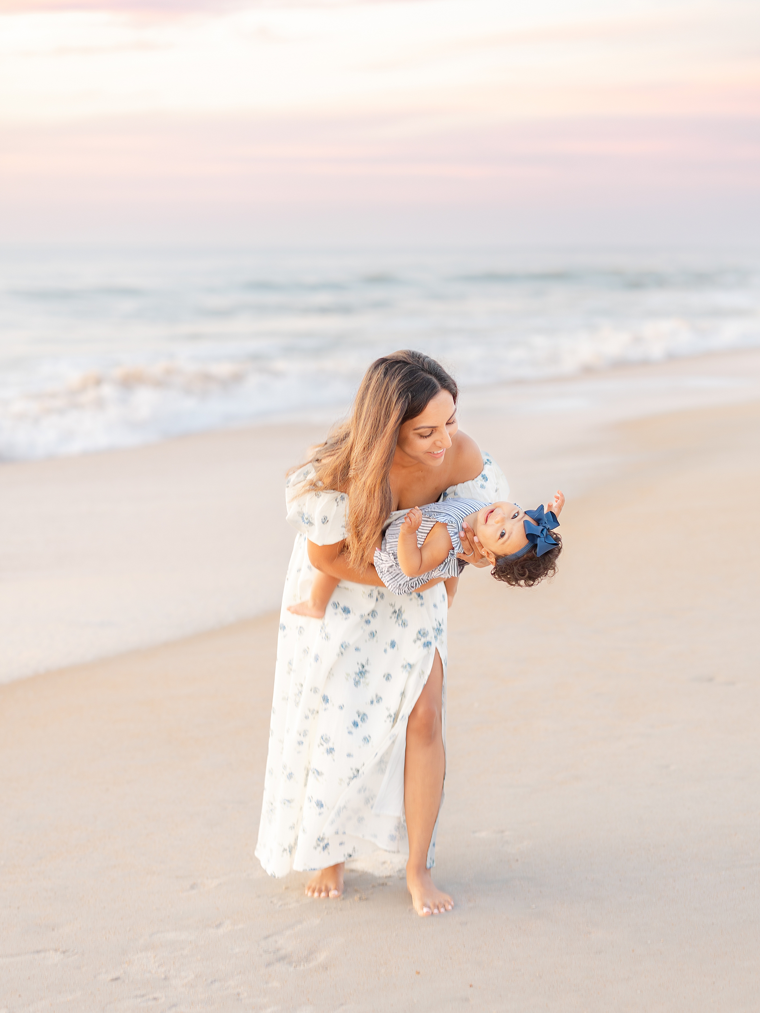 Woman in blue and white floral dress holding baby girl in striped romper on the beach at sunrise
