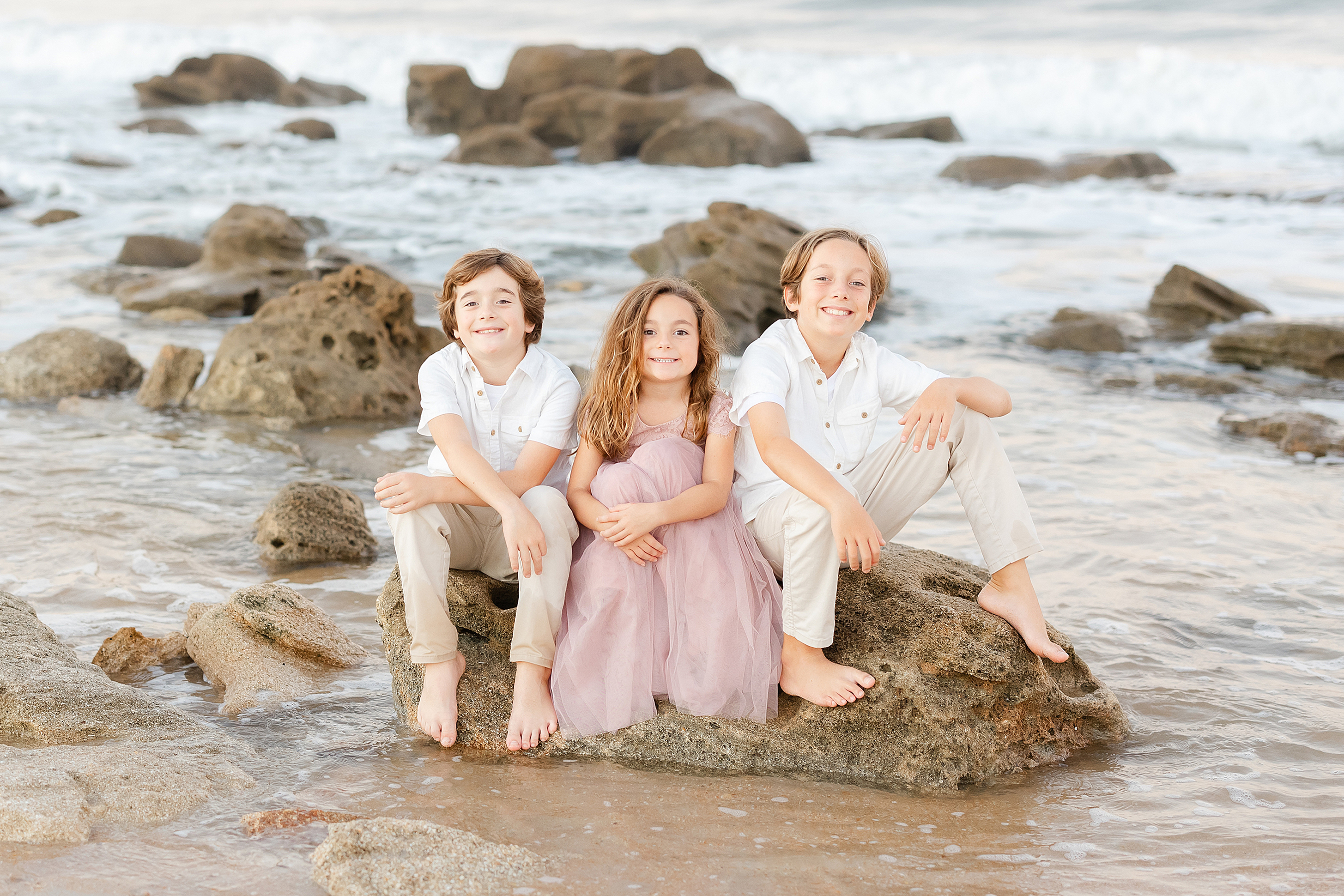 A sunset beach portrait of siblings sitting on the rocks together.