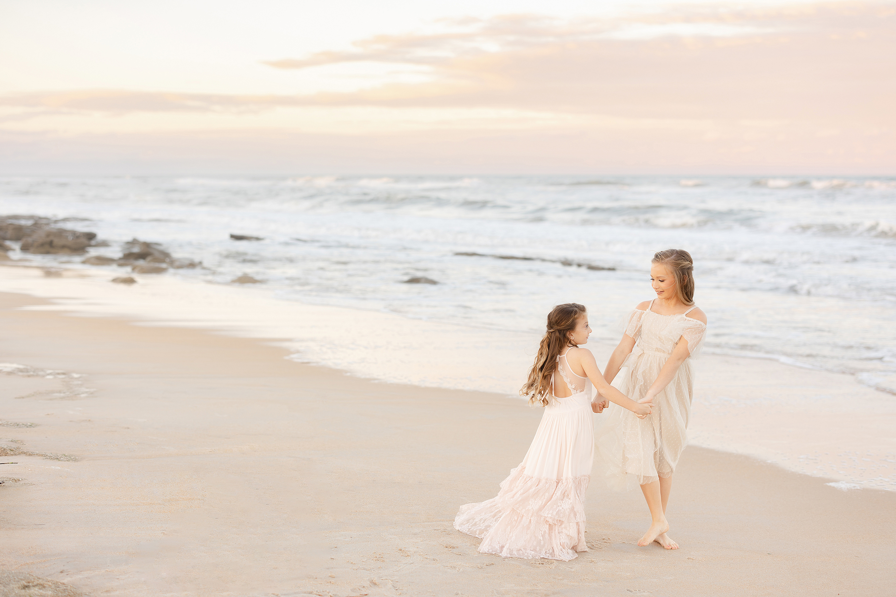 Two little girls dance together on the beach during a colorful pastel sunset.