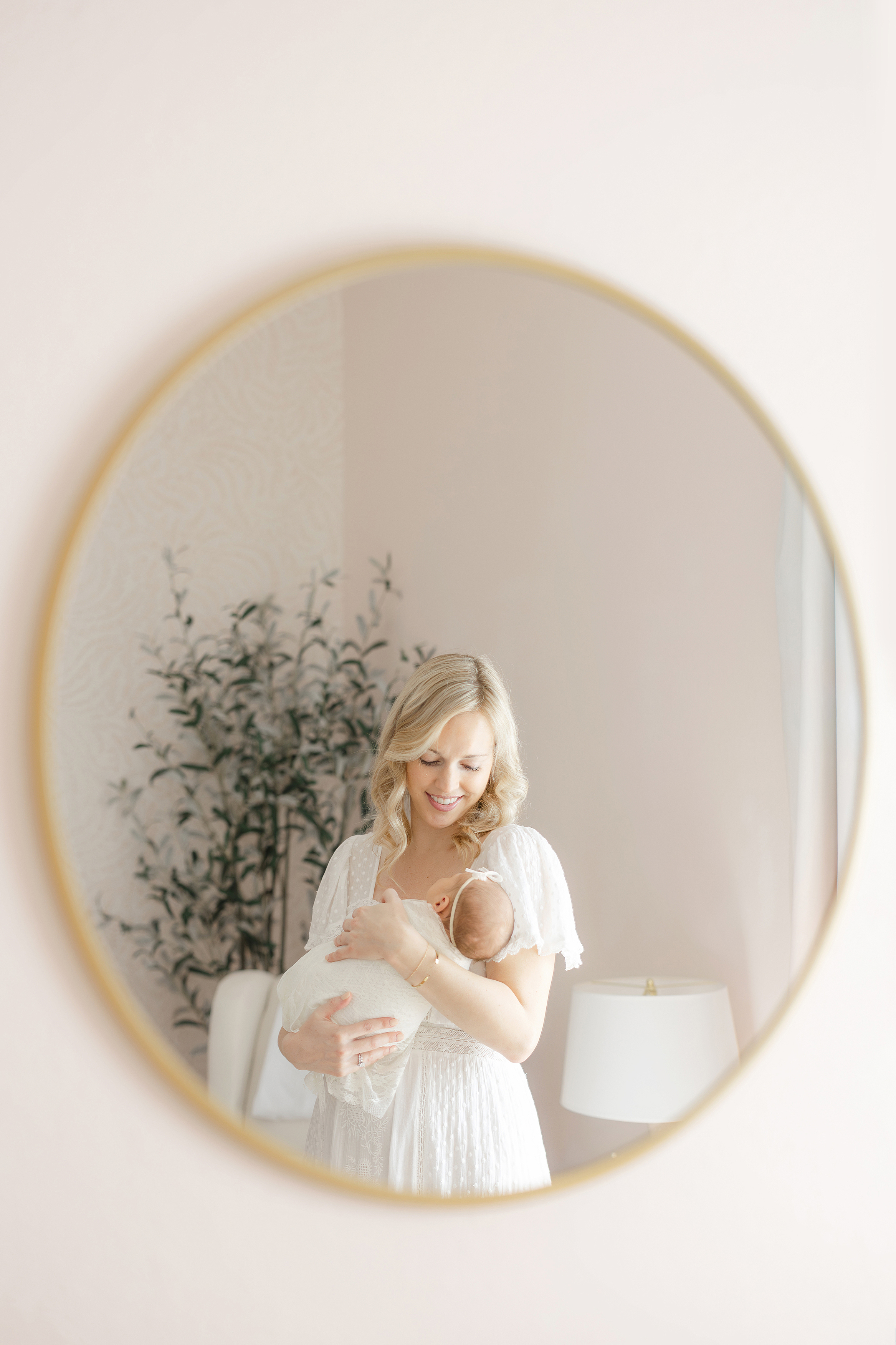 A blonde woman in a white lace dress holding a newborn baby girl in front of a mirror.