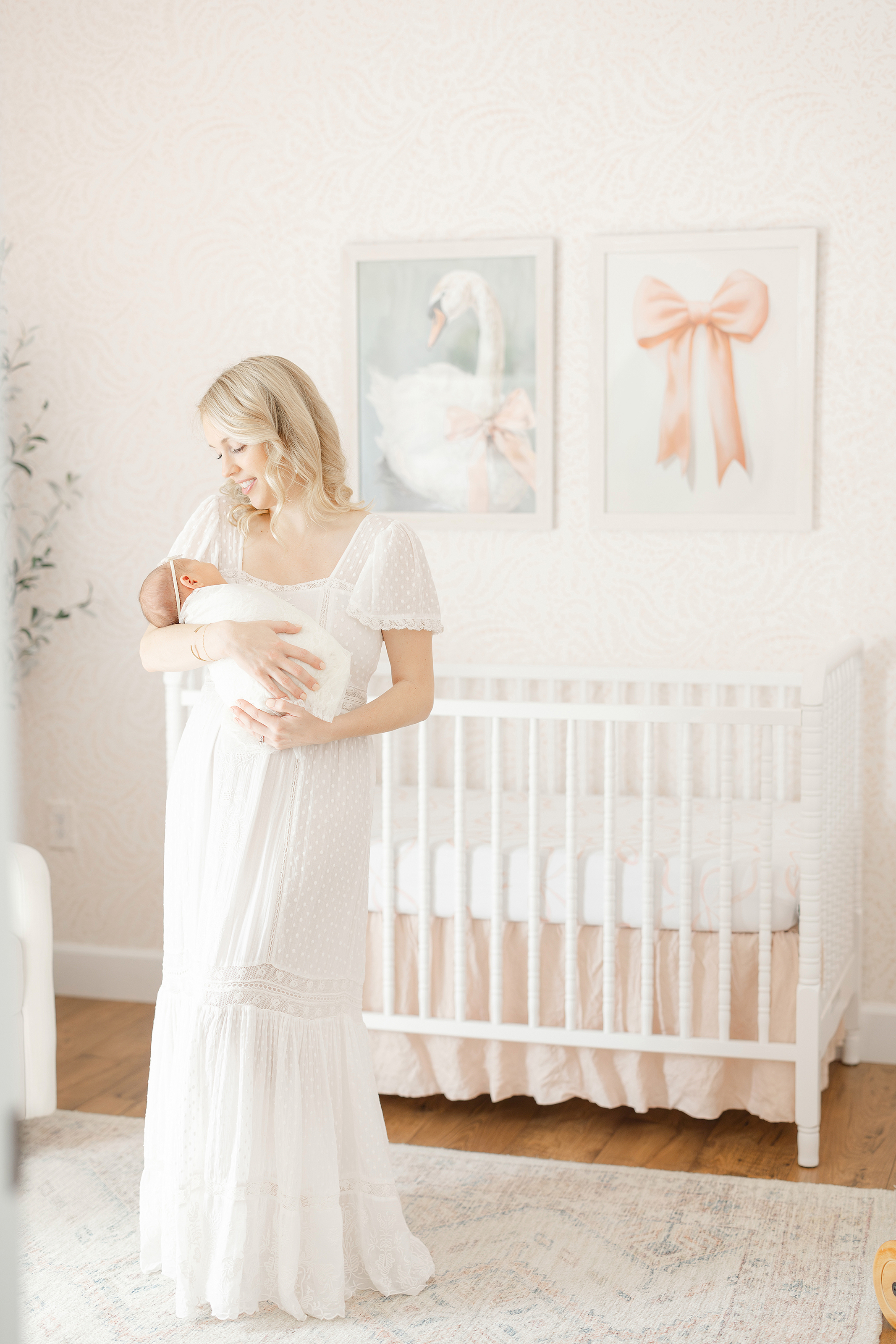 Blonde woman in white lace maxi dress holding a newborn baby girl in her nursery.