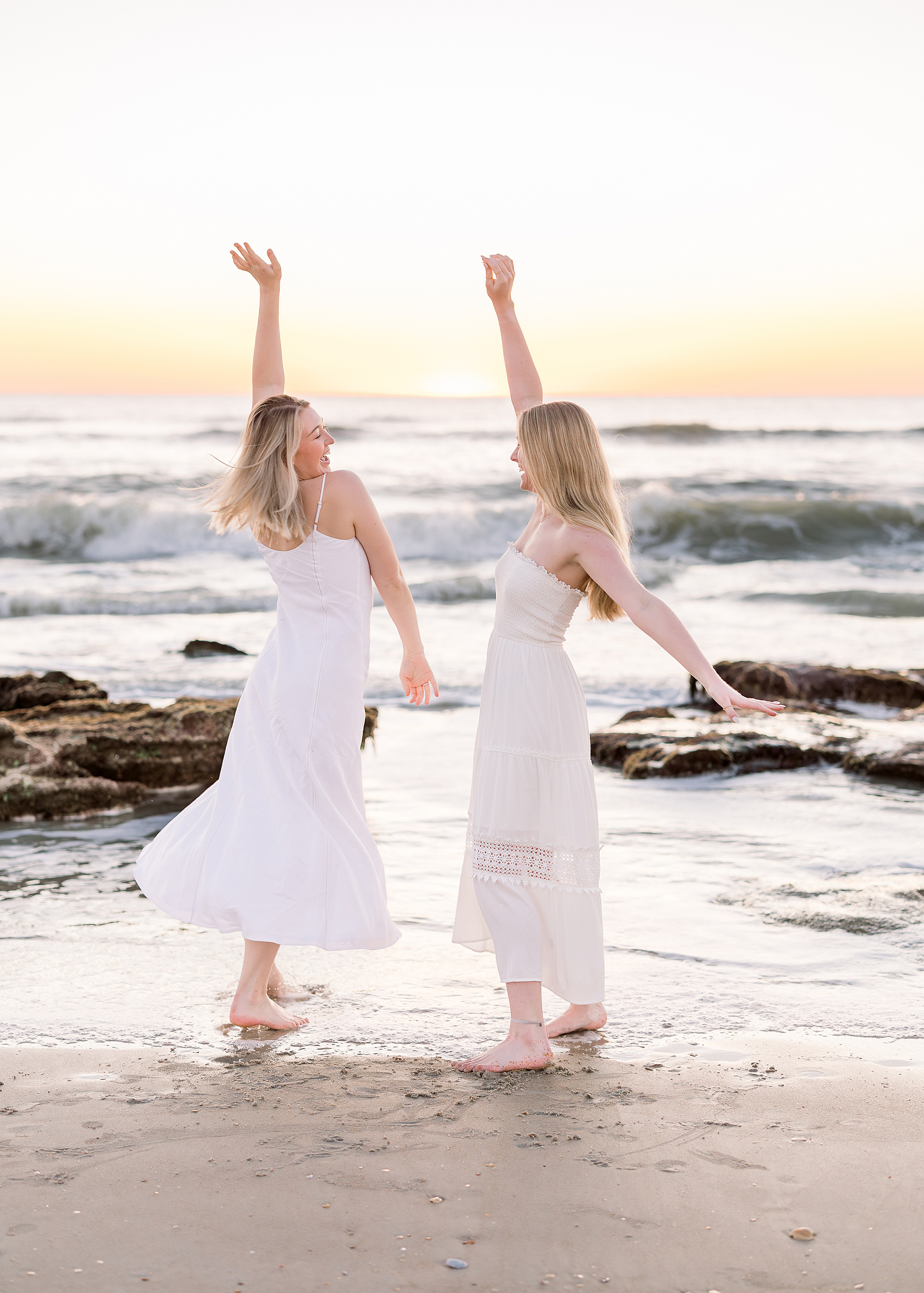 Two women in white dresses dancing at sunrise on the beach.