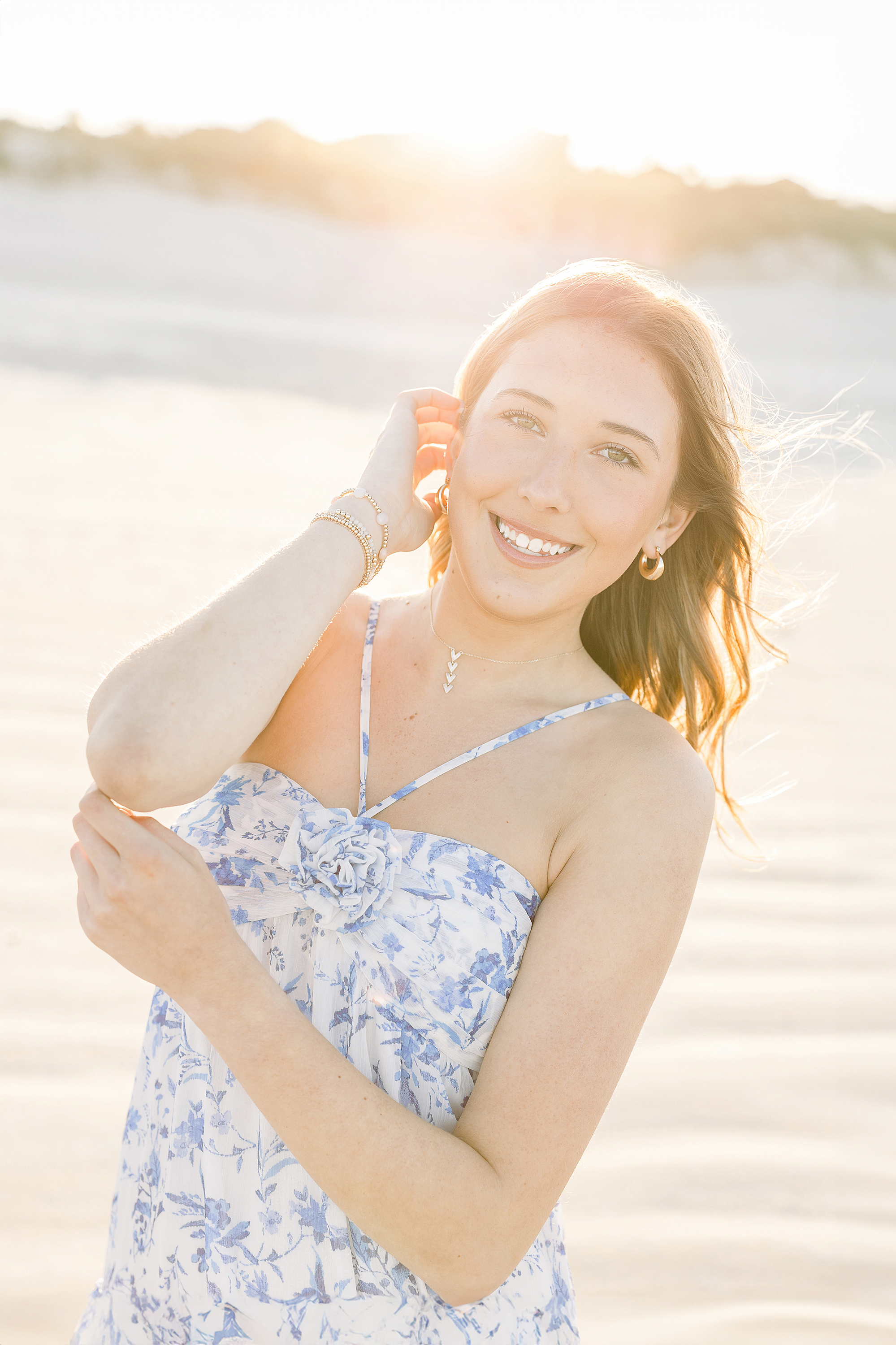 Backlit grad portrait of a young woman in a blue floral dress on the beach at sunset.