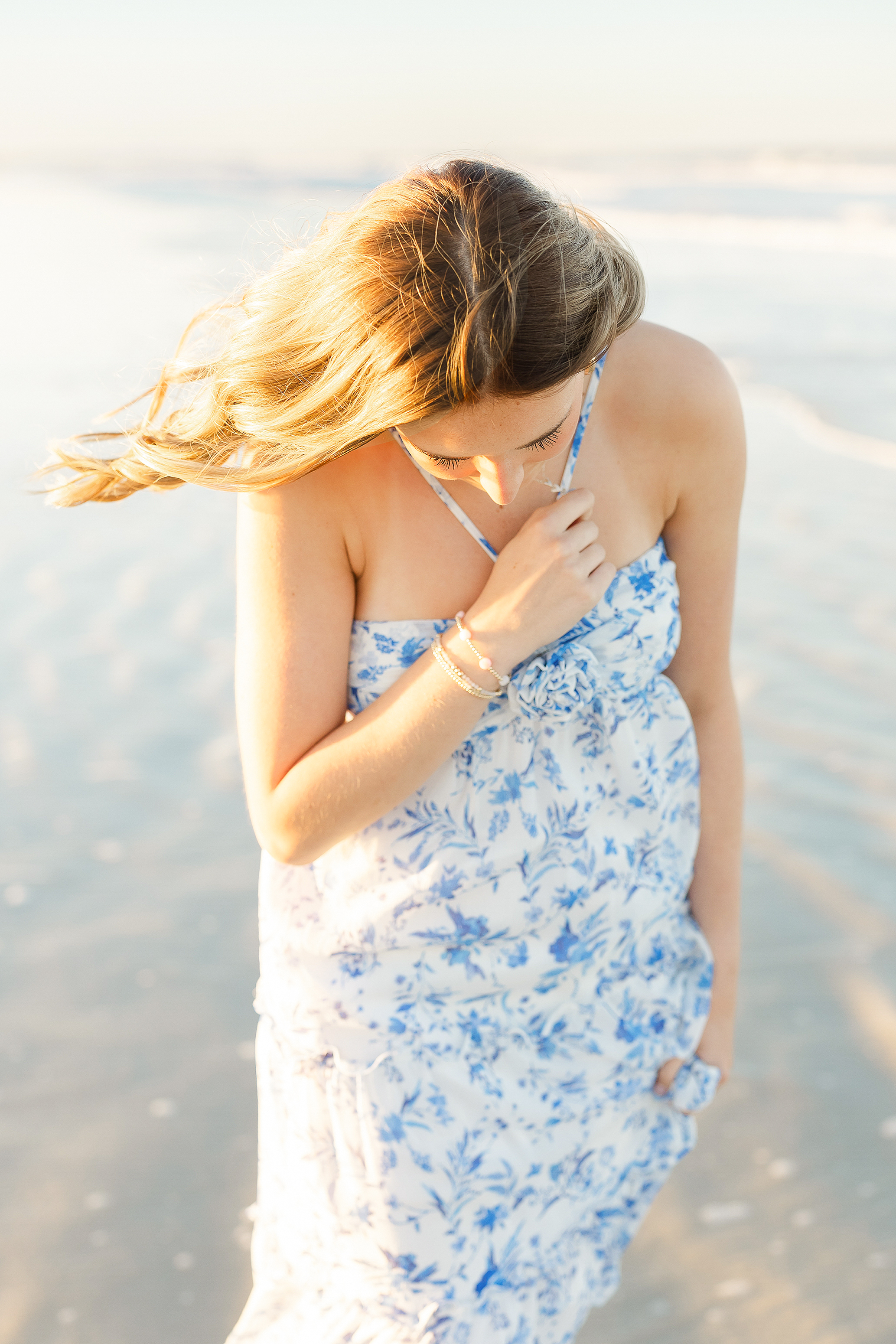 A young woman holds her necklace in warm sunlight on the sand.