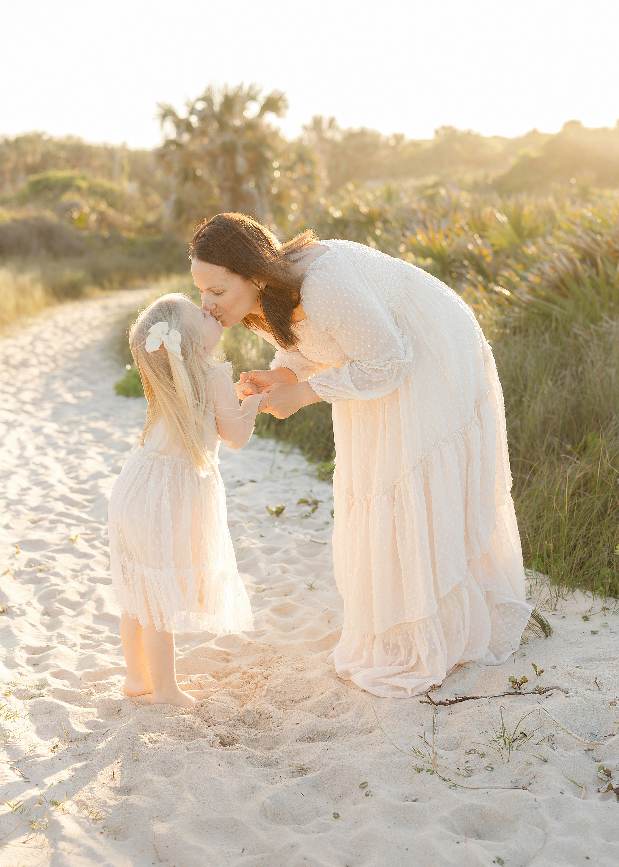 A mother dressed in cream tulle dress kisses her little girl in the backlit sun on the beach.