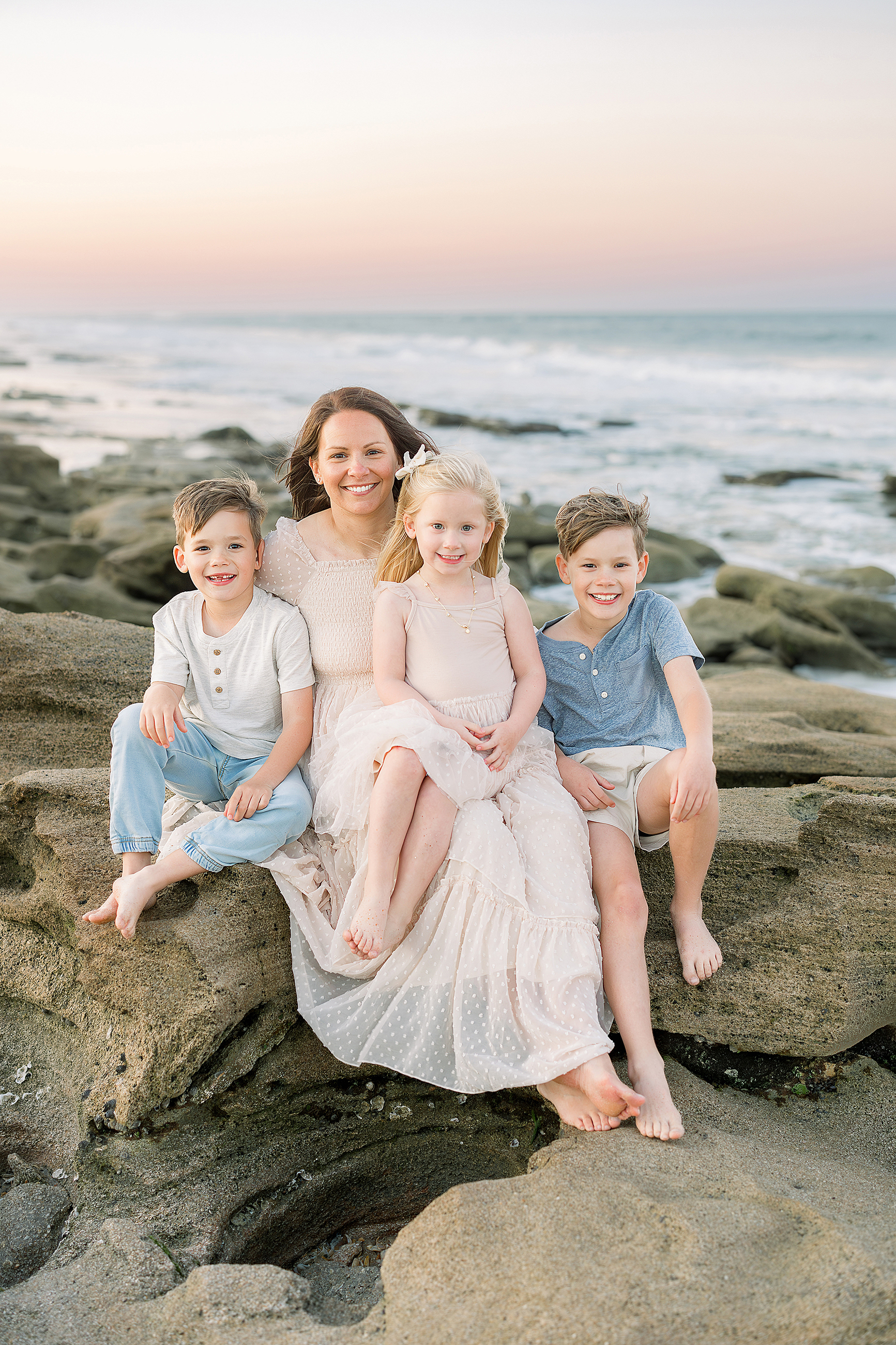 A mother sits on the rocks on St. Augustine Beach at sunset with her three little children.