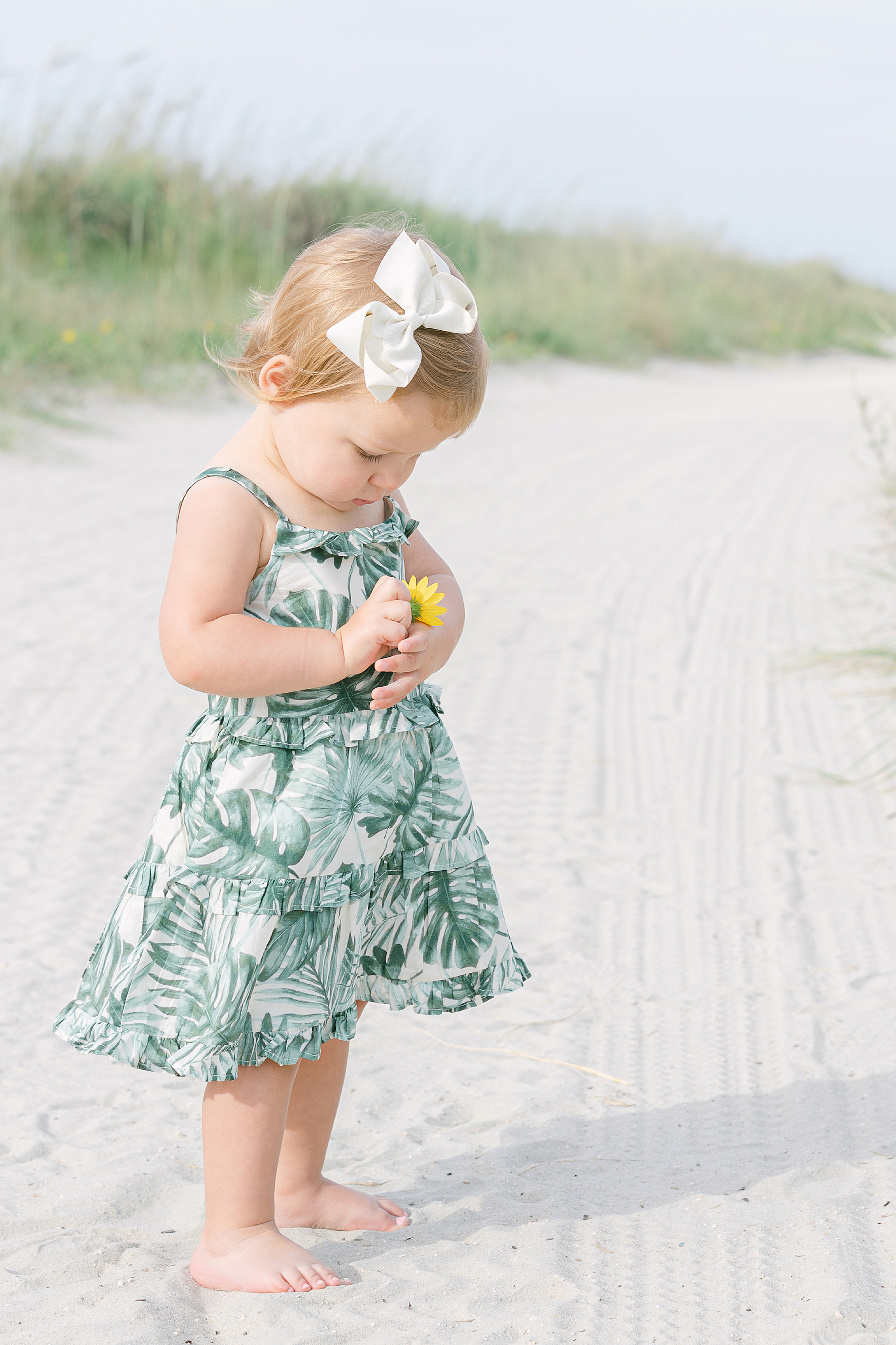 A toddler girl stands on the beach in Sea Island, GA holding a yellow flower.