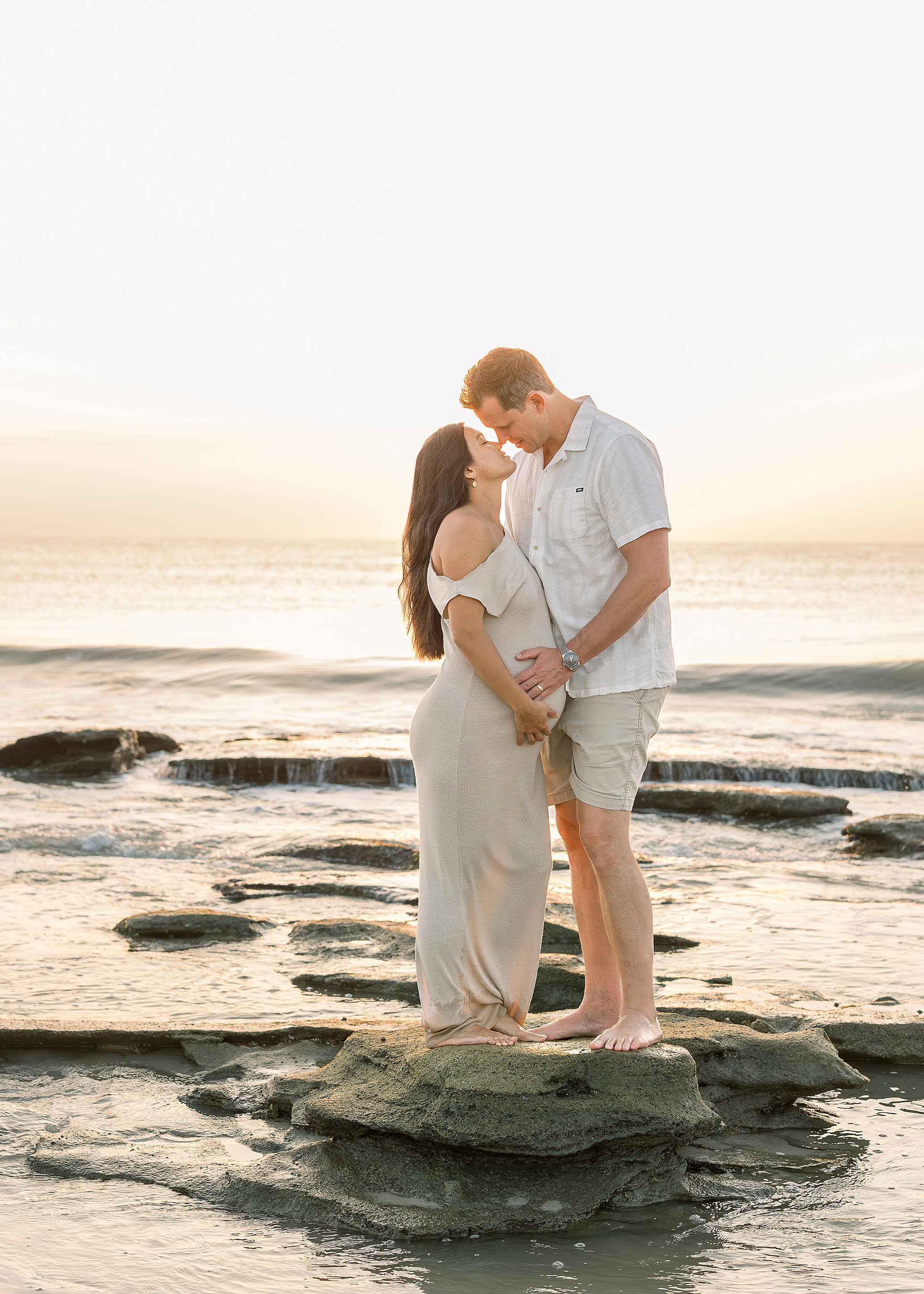 A couple stands on the rocks at sunrise on the beach embracing each other.