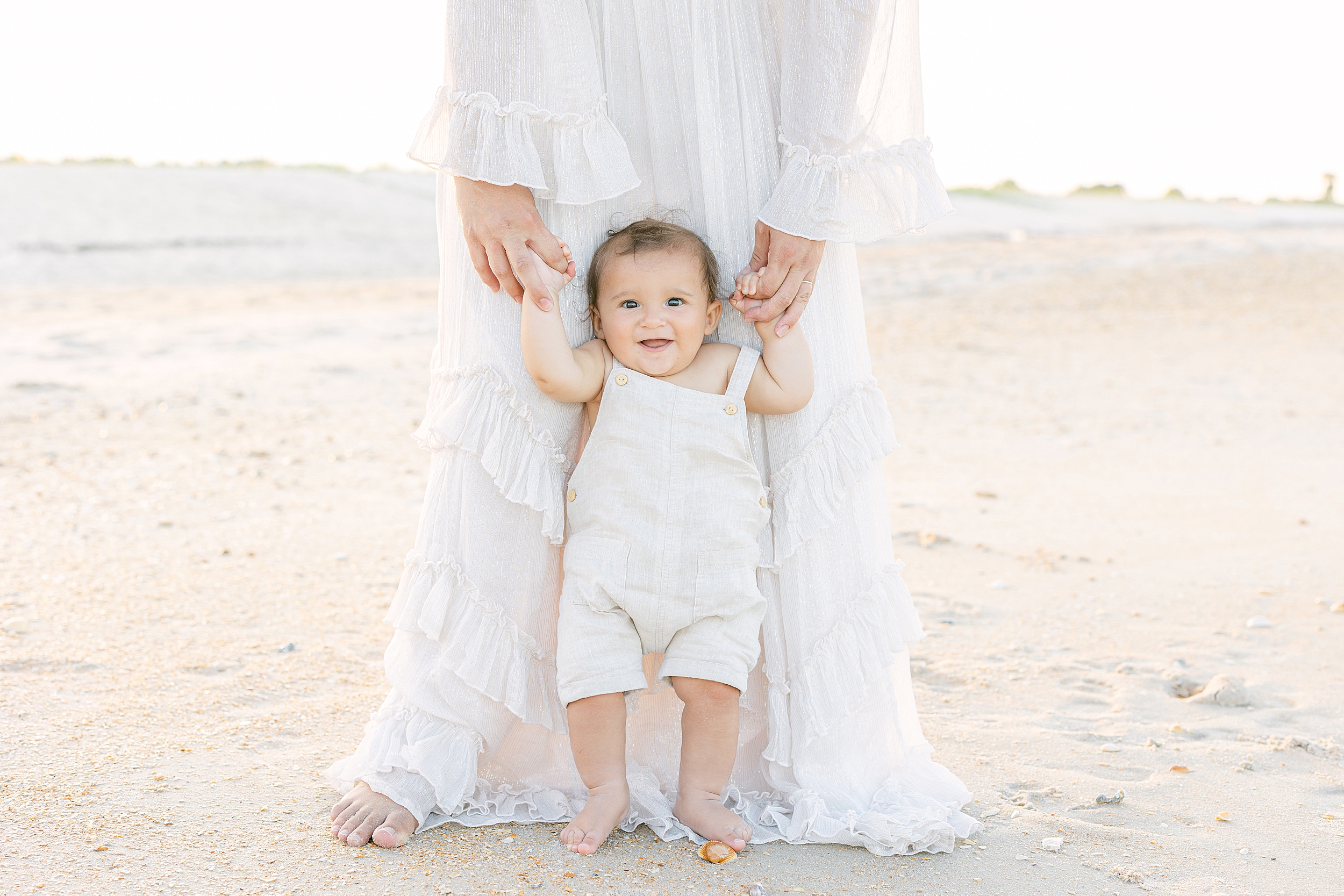 A baby boy dressed in neutral linen overalls stands next to his mother on the beach at sunset.