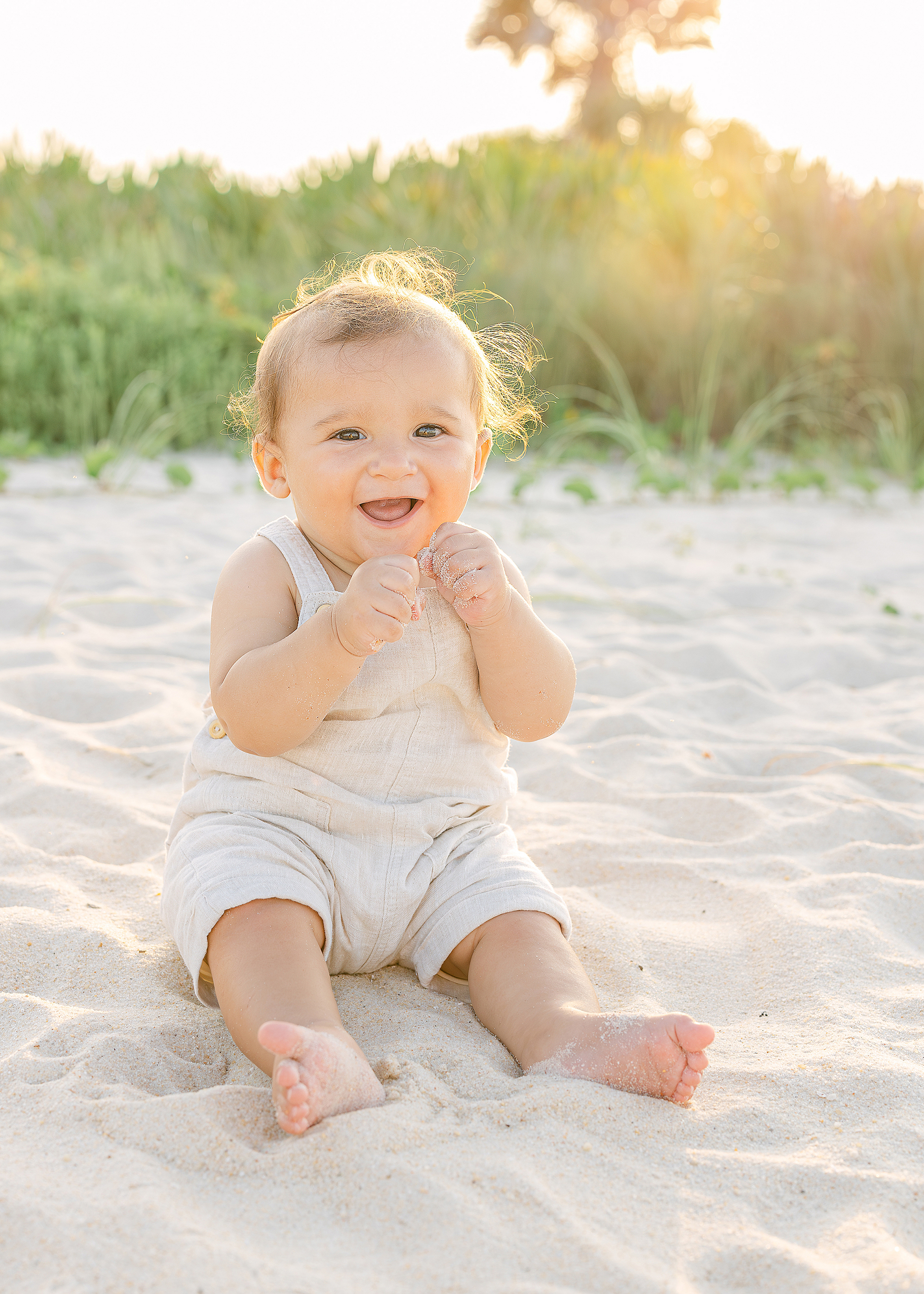 A baby boy in a tan linen onsie sits on St. Augustine Beach at sunset.