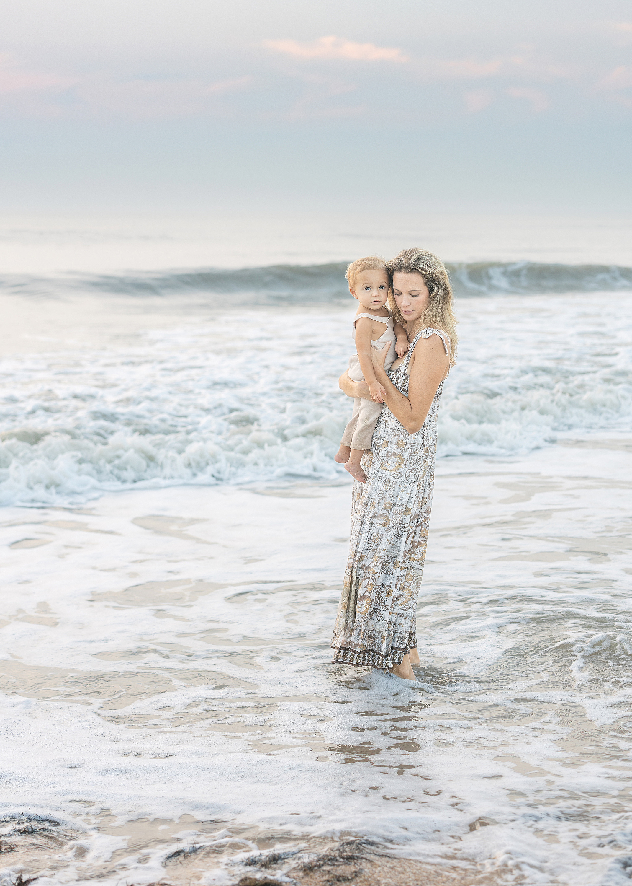 A woman holds her baby boy in the water on the beach at sunrise.