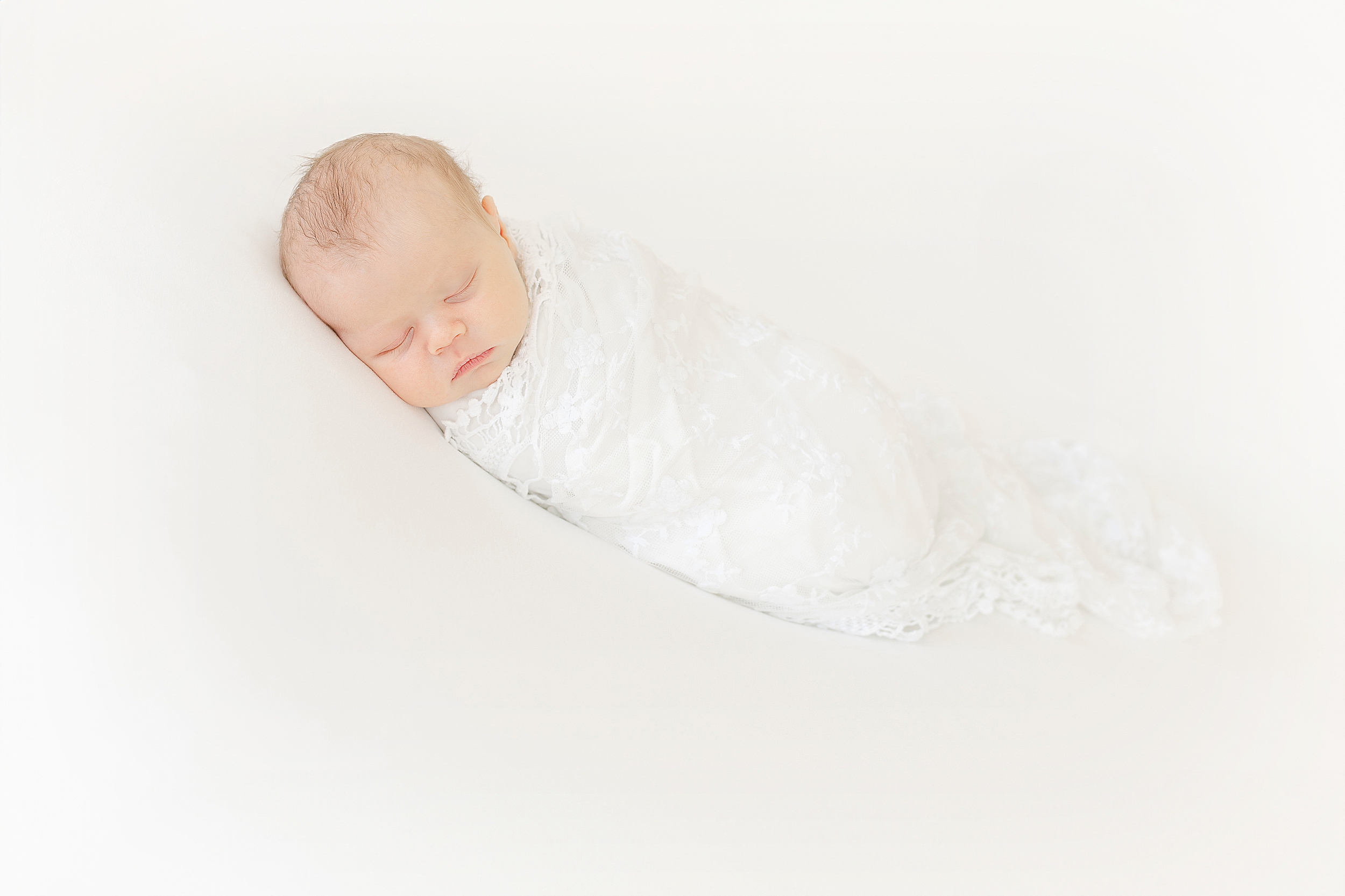 An all white newborn portrait of a newborn baby girl wrapped in a white lace swaddle on a white blanket.