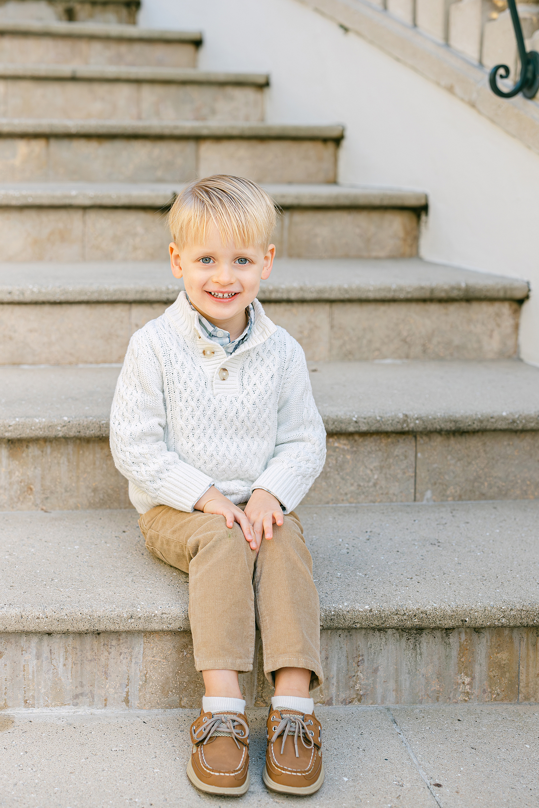 A little blonde haired boy dressed in neutrals sits on the steps of The Cloister.