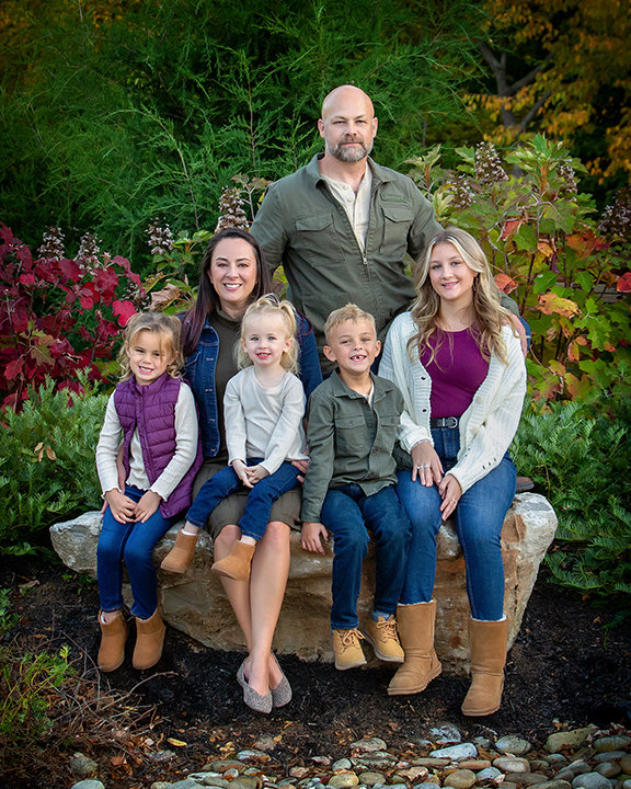 Family of six posing outdoors with greenery and colorful foliage in the background.