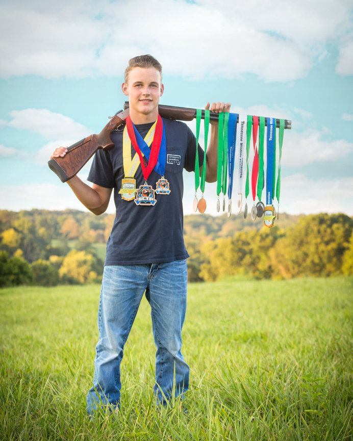 A person standing in a field holding a shotgun and displaying numerous medals on their arm.