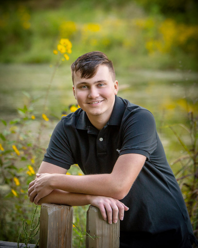 Young man in a black polo leaning on a wooden fence with a field of yellow flowers in the background.