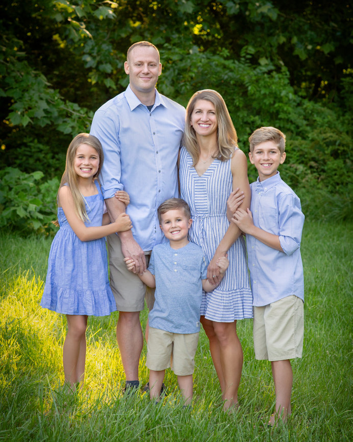 Family of five smiling in a grassy area with trees, all wearing blue and white summer outfits.
