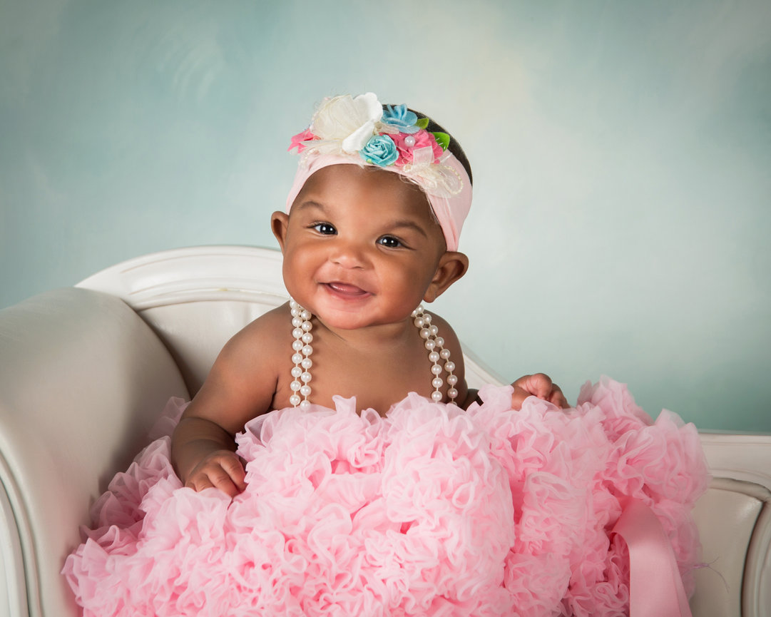 Smiling baby wearing a pink frilly dress and floral headband, sitting on a white chair against a soft background.