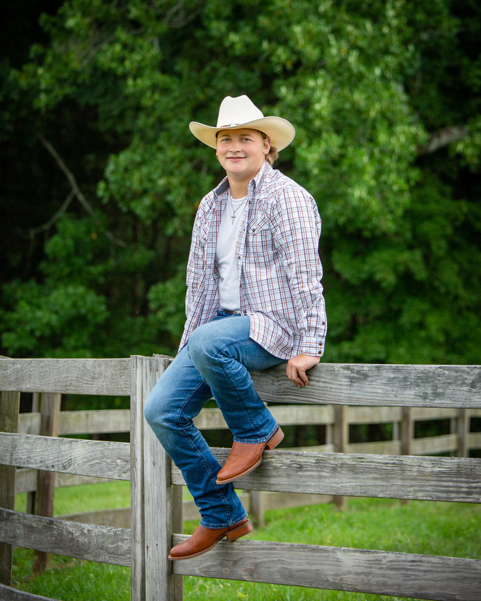 Man in cowboy hat and plaid shirt sits on a wooden fence, with lush green trees in the background.