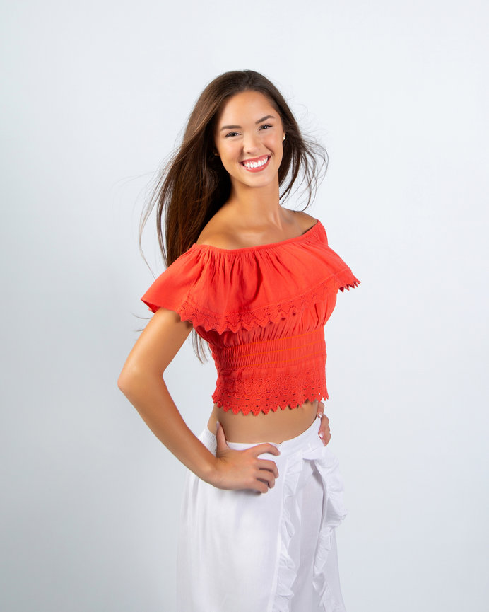 Smiling woman in a red off-shoulder top and white pants, standing against a plain background.