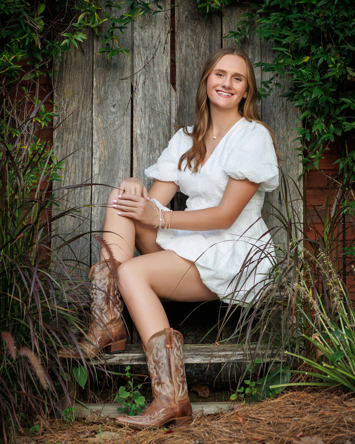 A woman in a white dress and cowboy boots sits on a wooden bench surrounded by greenery, smiling gently.