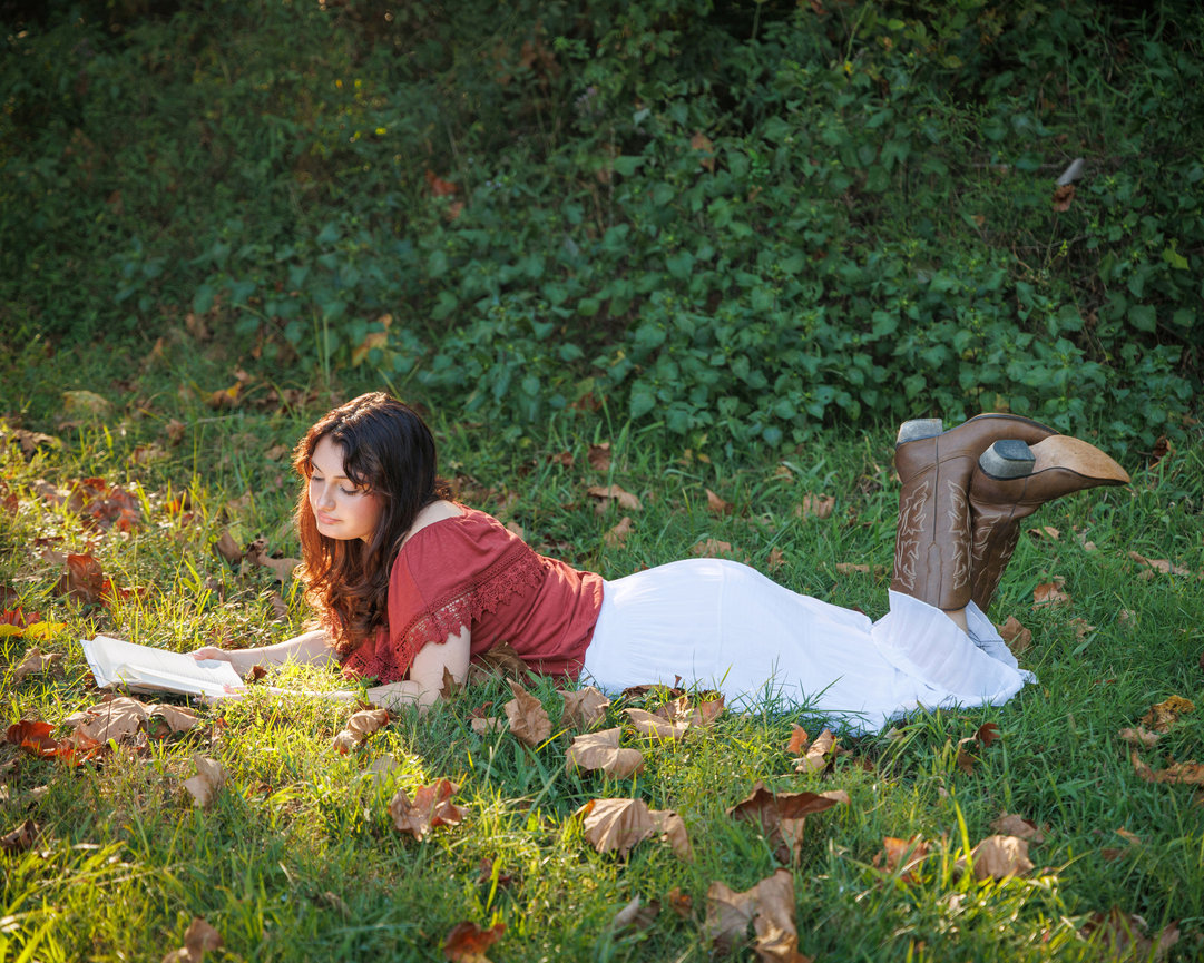 Woman reading a book on grass with autumn leaves, wearing a red blouse, white skirt, and cowboy boots.