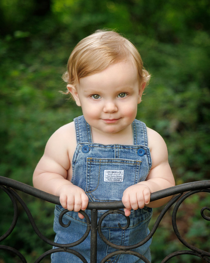 A toddler with curly hair in denim overalls leans on a black metal fence, surrounded by lush greenery.