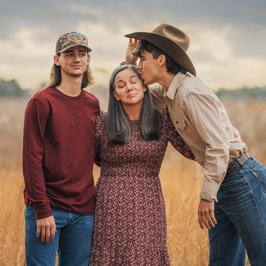 Three people pose playfully in a field. A woman in the center with a patterned dress, flanked by two men, one in a cowboy hat.