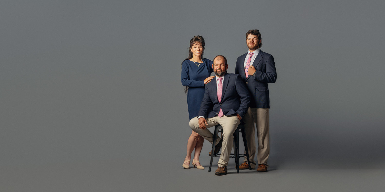 Three people in formal attire, posing against a gray background. One person is seated on a stool, flanked by two standing.