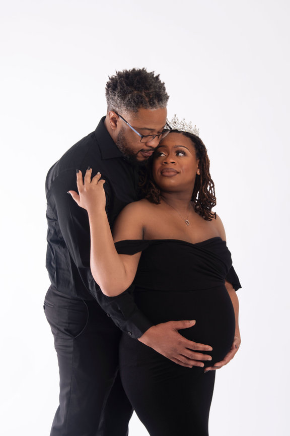 Expecting couple joyfully posed, with the woman in a black off-shoulder dress and tiara, cradling her baby bump.