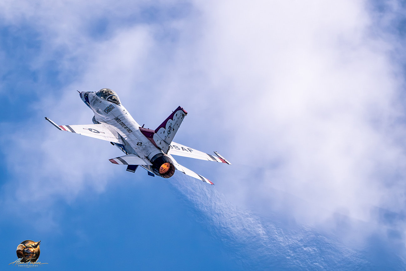 U.S. Air Force Thunderbirds F-16 Fighting Falcon climbing steeply with orange afterburner visible against a cloudy blue sky.