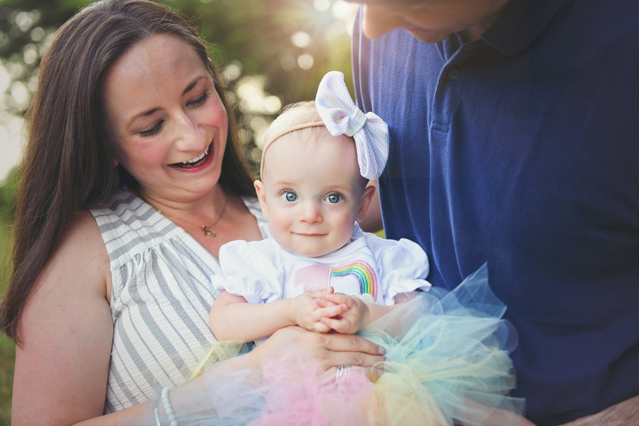 Mother and father holding their baby girl wearing a rainbow tutu and bow during a milestone first birthday session in Mocksville, NC by Megan Gioeli Photography