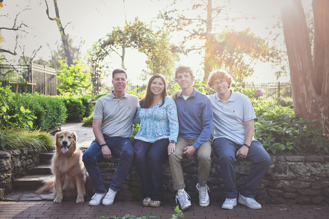 Family of four with teenage sons and their golden retriever sitting on a stone bench in a lush garden at sunset, photographed in Clemmons, NC by Megan Gioeli