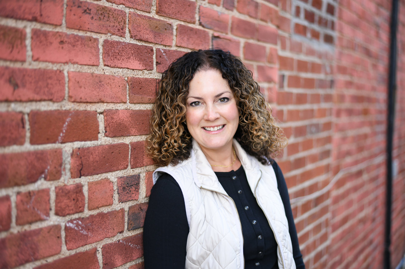 Woman with curly hair smiling and standing against a red brick wall for a professional headshot.