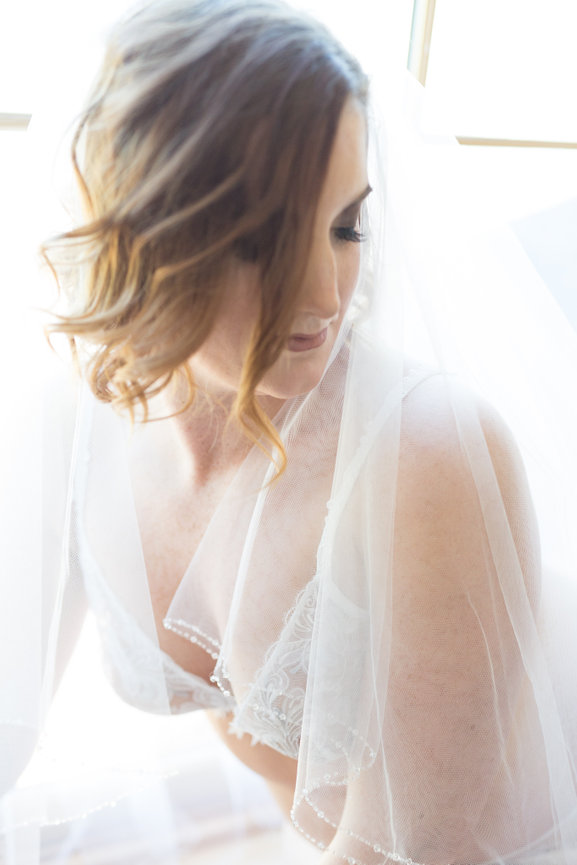 Bride in a white lace dress and veil, looking down softly, illuminated by natural light.
