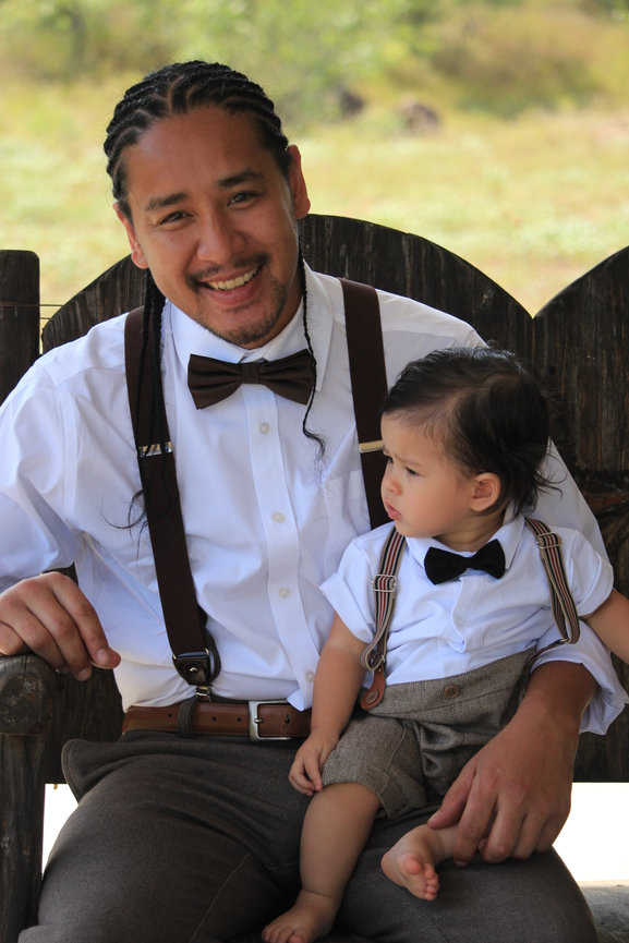 Father in a bow tie and suspenders smiling with his baby son in a matching outfit, outdoor family portrait