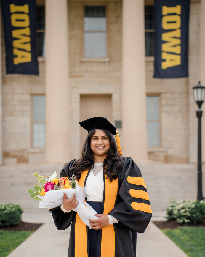 Graduate in cap and gown holding a bouquet of flowers, smiling in front of Iowa university building with flags, Iowa City.