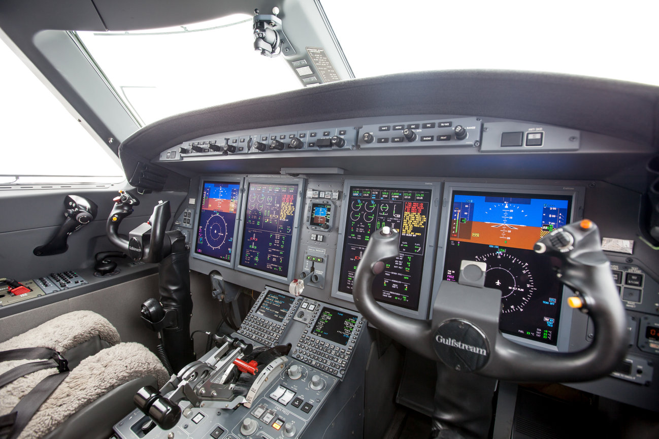 Cockpit interior of an aircraft with multiple digital displays, controls, and two yokes.