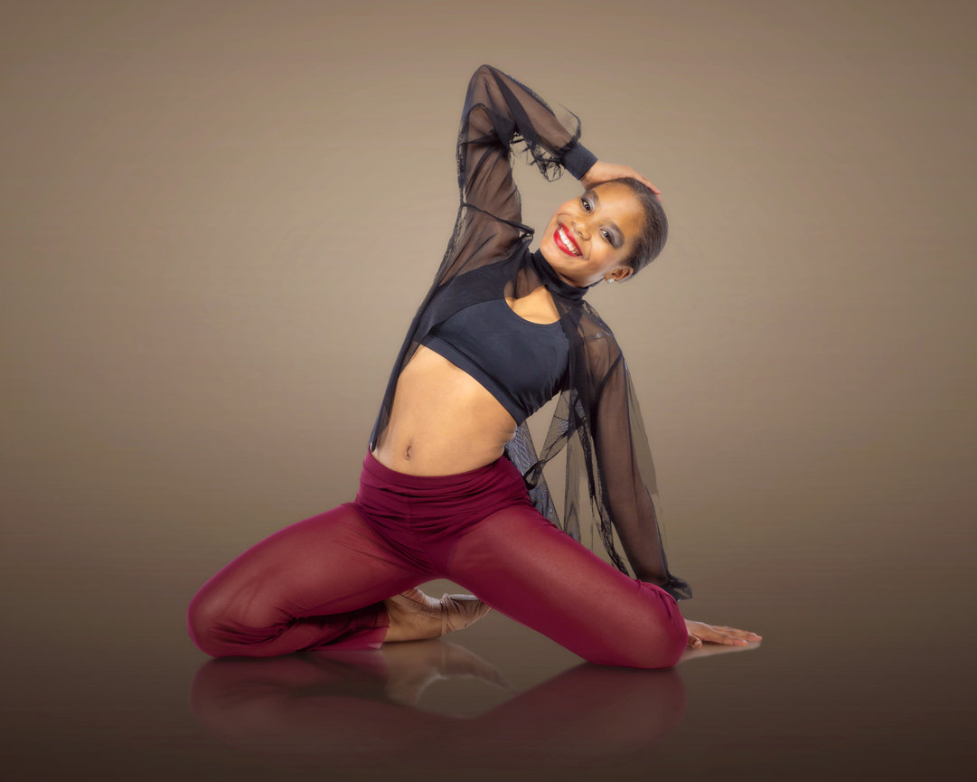 Person kneeling on one knee, wearing a black top and sheer burgundy pants, smiling with one hand on head, against a gradient background.