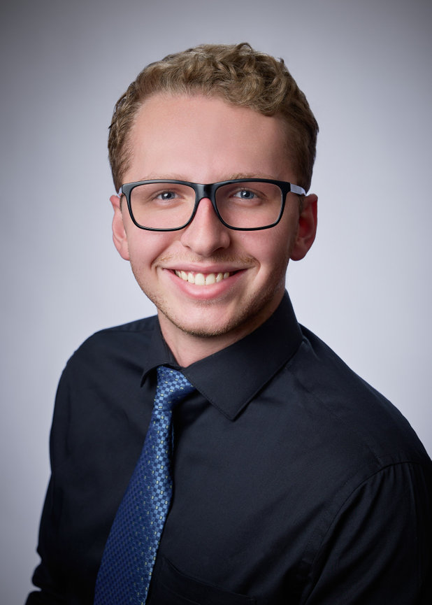 Headshot of a smiling young man in glasses wearing a black shirt and blue tie against a gray background