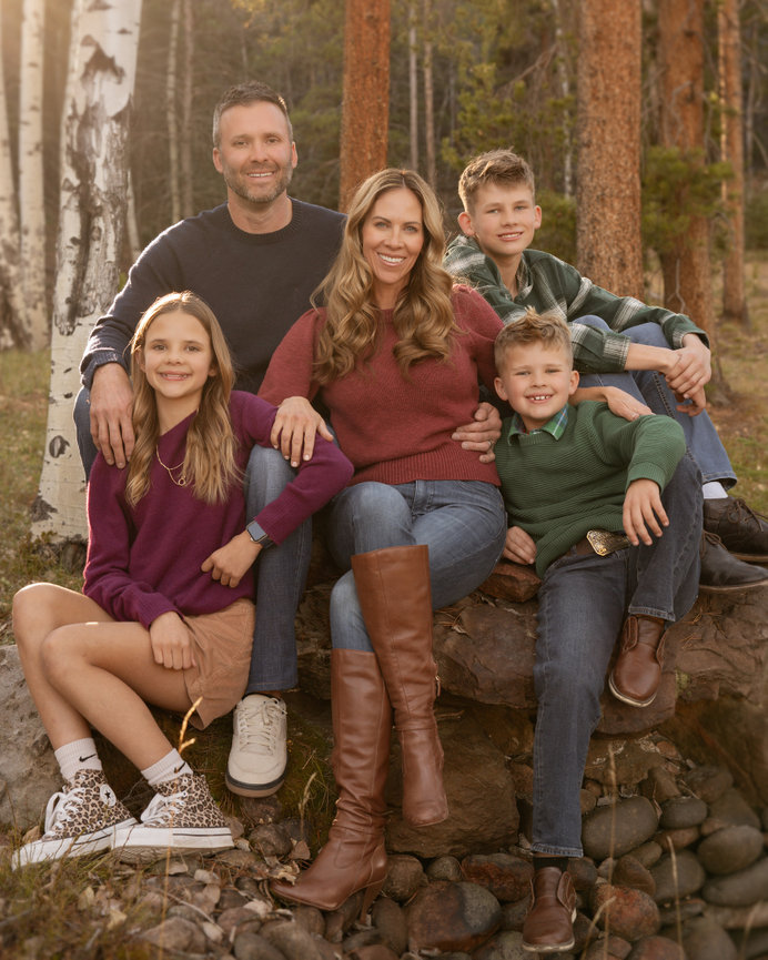 A family of five sitting outdoors on rocks, surrounded by trees; everyone is smiling.