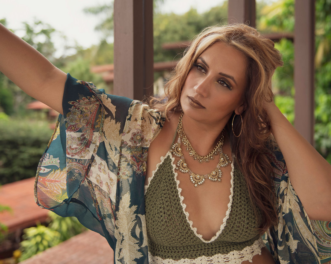 A woman poses outdoors wearing a floral kimono, green crochet top, and layered necklaces, surrounded by greenery.