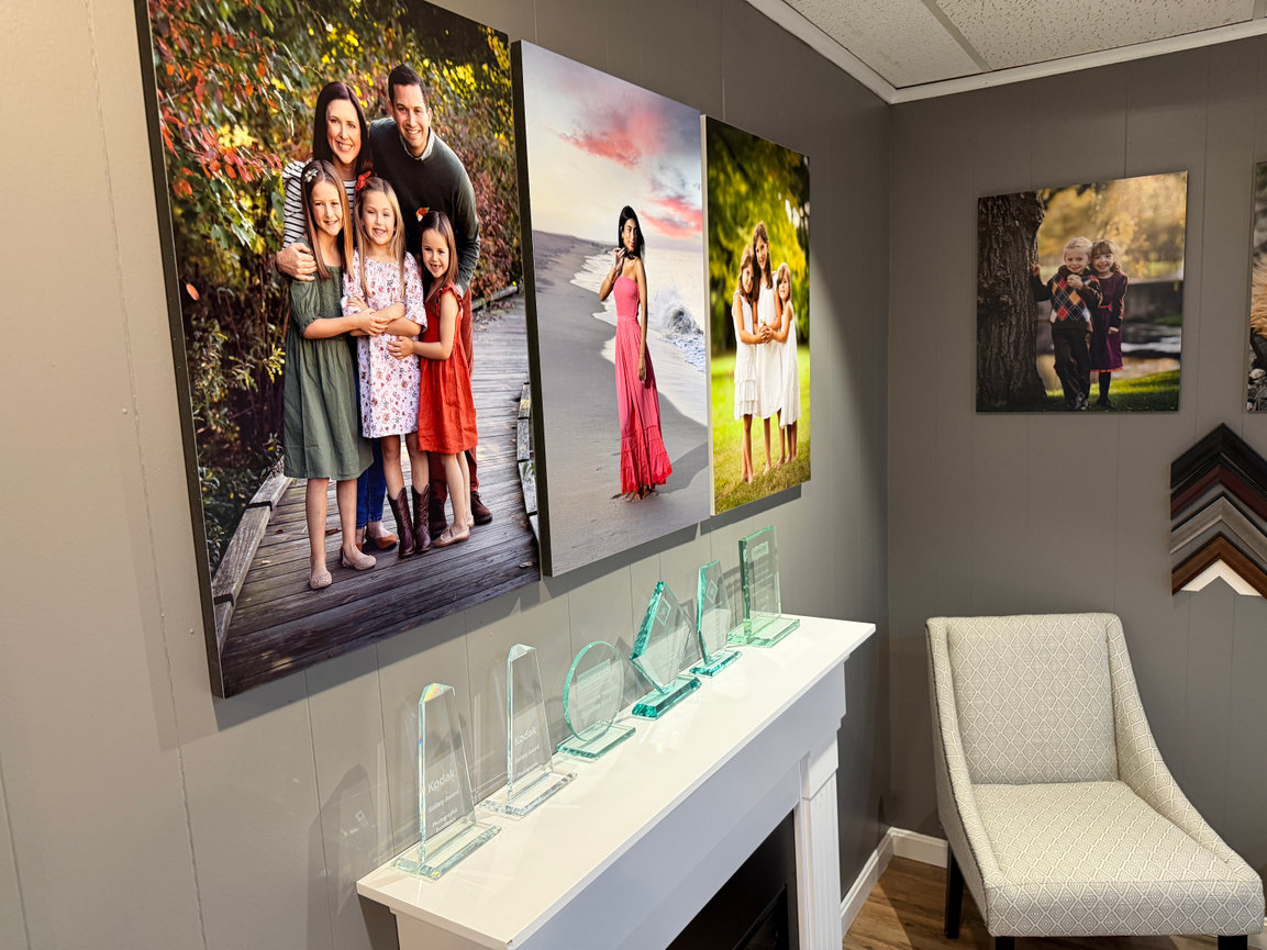 Family portraits and awards displayed on a wall above a white fireplace in a cozy gallery room at Paula Swift Photography studio in Metrowest Boston.