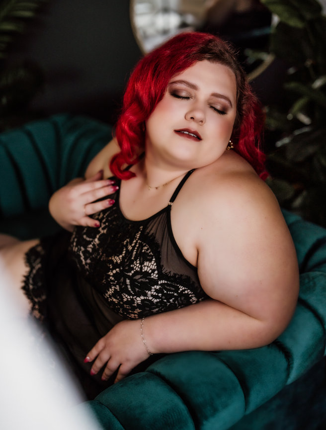 amy matney boudoir studio port angels, wa sexy curvy boudoir Person with red hair wearing black lace clothing, reclining on a teal tufted chair with relaxed expression.