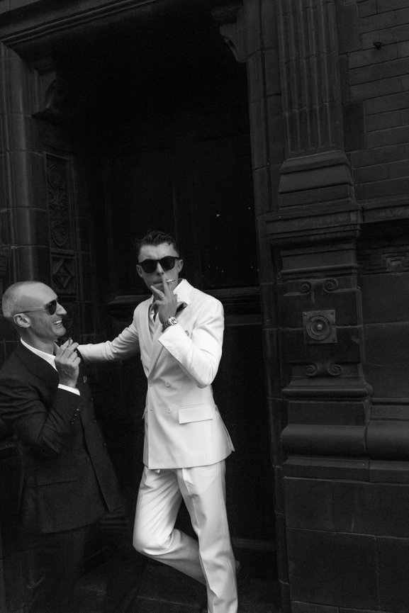 Editorial Manchester wedding photography: two grooms in suits and sunglasses posing against an ornate dark building facade.