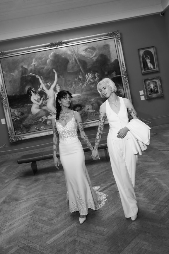Editorial wedding photography of two brides holding hands at Manchester Art Gallery, walking past a grand classical painting in a modern, cinematic style.