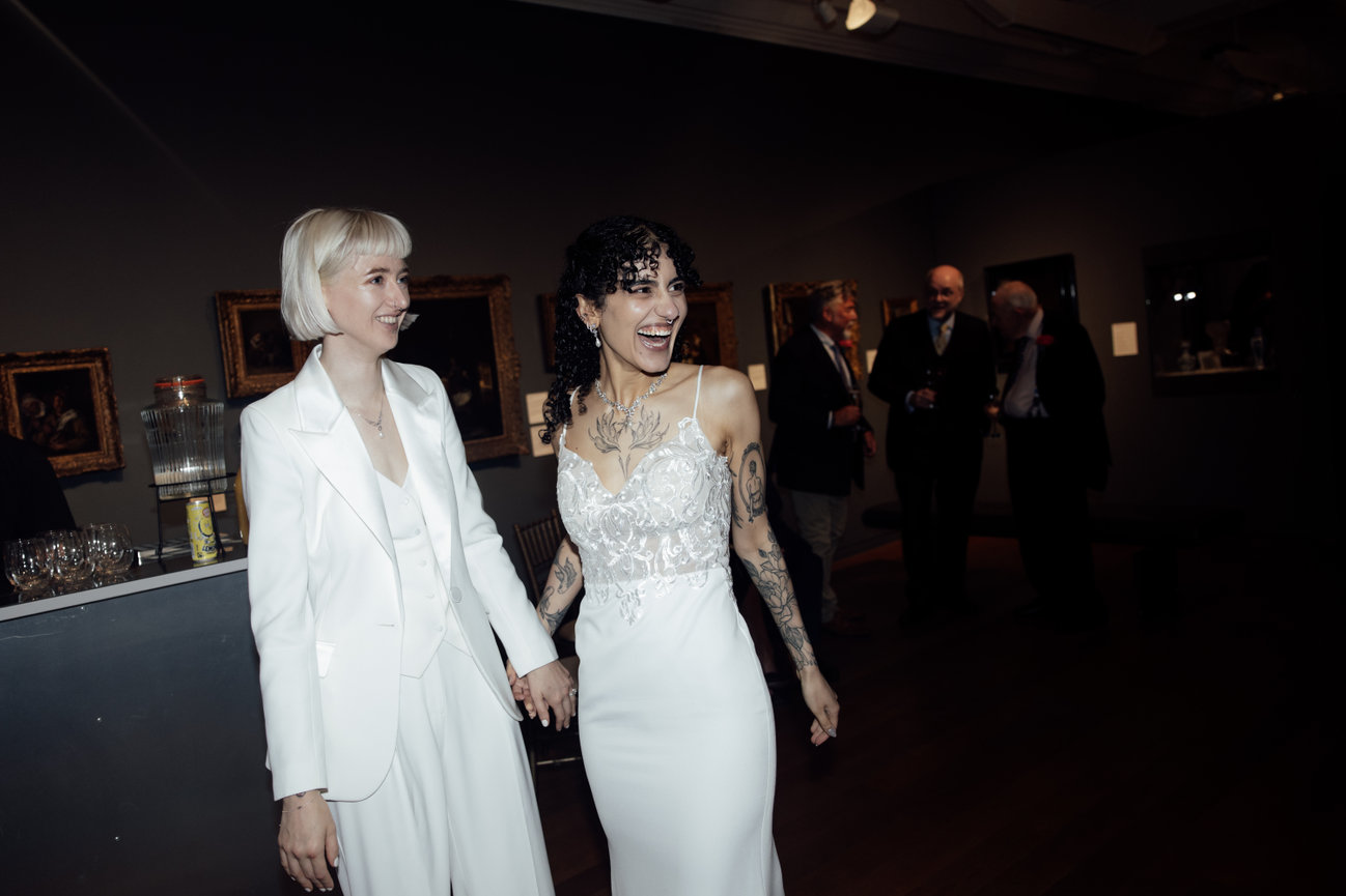 Direct flash editorial photography of two smiling brides—one in a modern white trouser suit and the other in a lace wedding dress—holding hands amidst guests and classical paintings at Manchester Art Gallery.