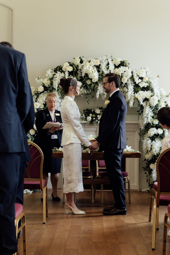 St George's Hall, Liverpool: A couple stands holding hands during a wedding ceremony with a flower arch backdrop.