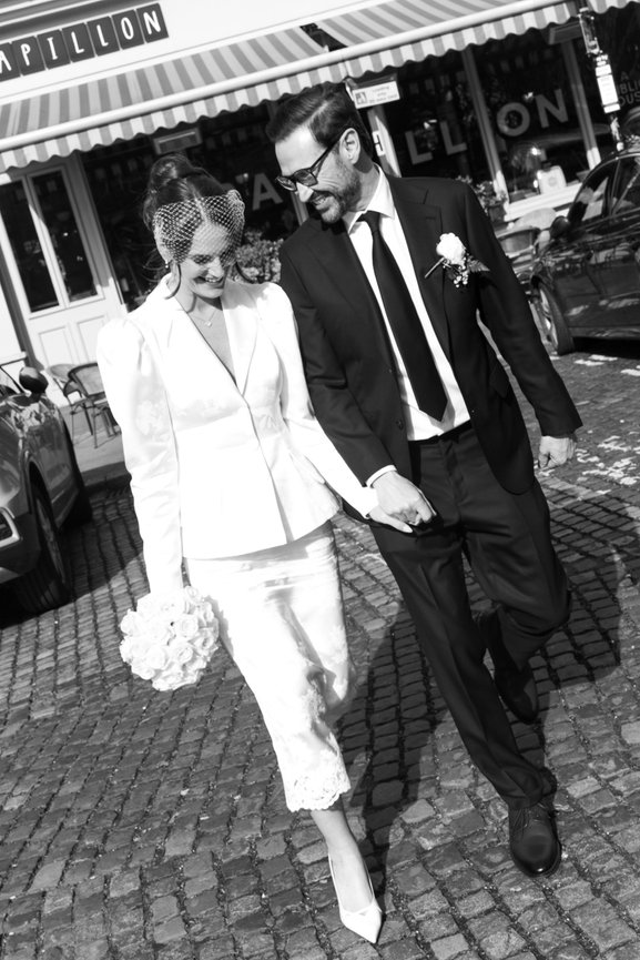 A bride & groom in wedding attire walks hand in hand on a cobblestone street in Liverpool, captured in black and white, documentary feel.