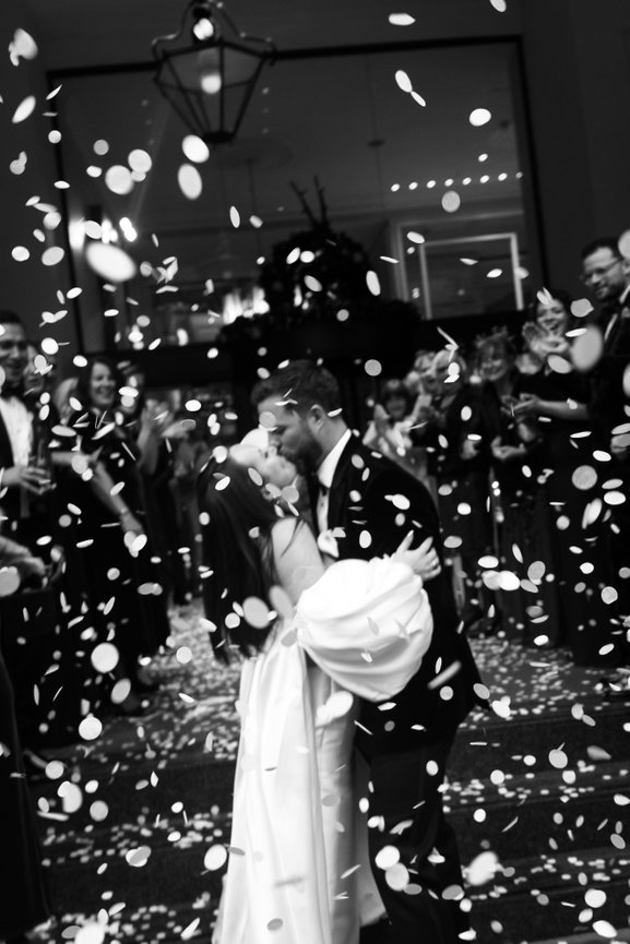 High-contrast monochrome wedding photography at The Midland Hotel, Manchester. Confetti motion of bride and groom kissing surrounded by celebrating guests.